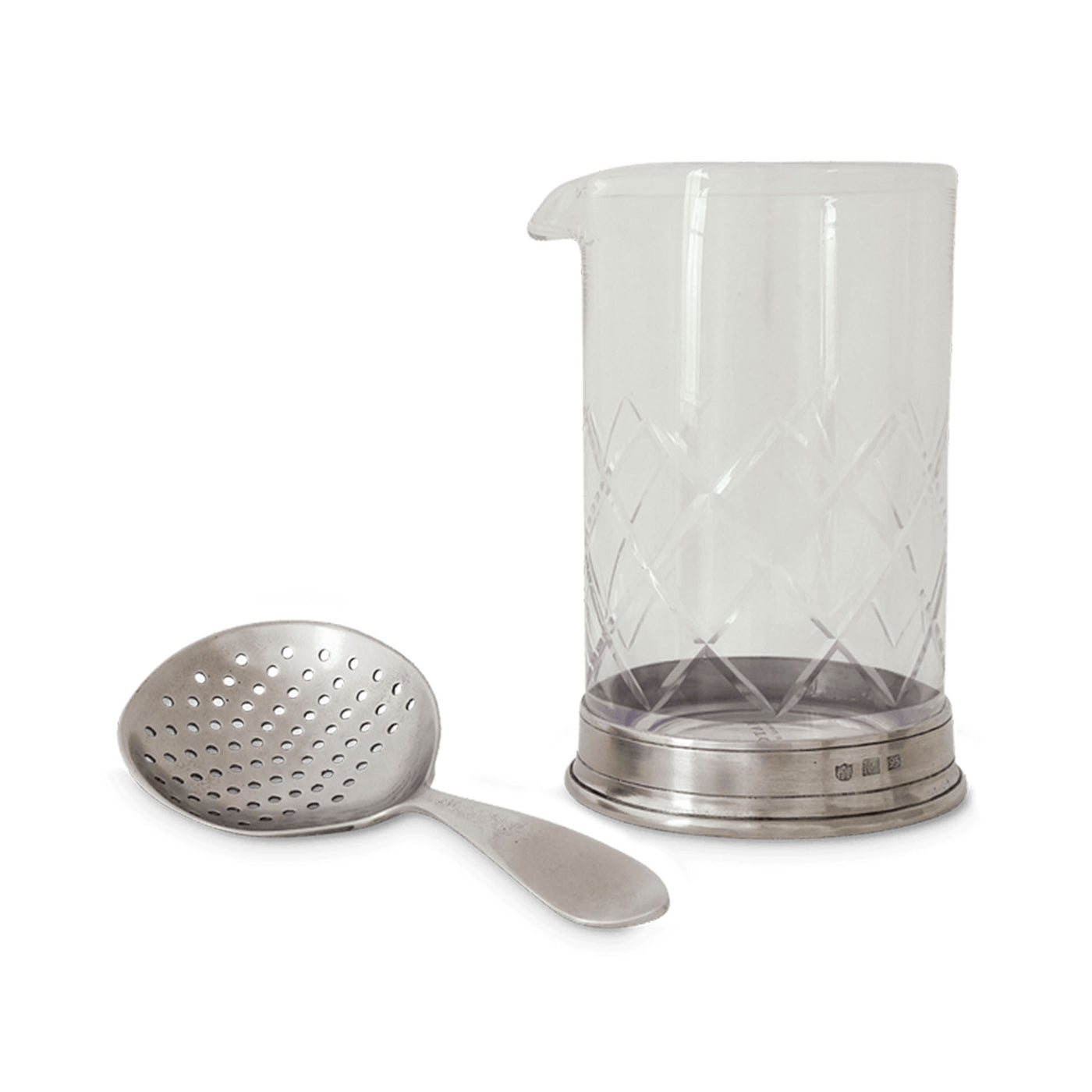 A clear mixing glass with a geometric pattern and a pewter cocktail strainer beside it.